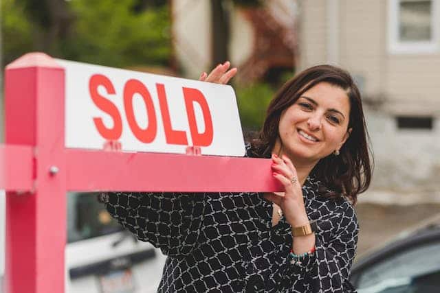 smiling-woman-standing-next-to-a-red-and-white-sold-real-estate-sign-in-front-of-a-house